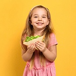 A little girl in a pink dress and headband smiling while holding a sandwich with lettuce on it
